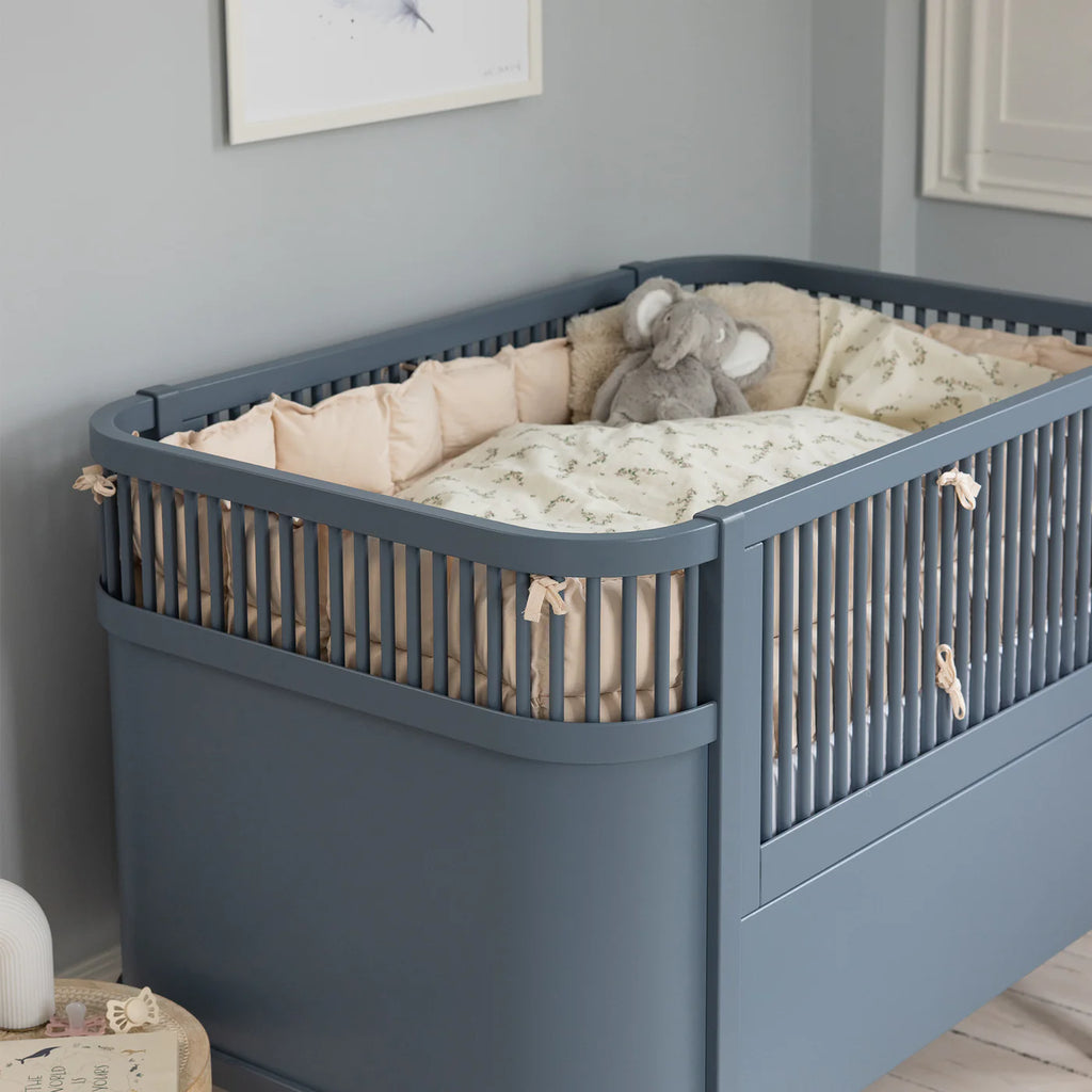 Blue crib with beige bedding and a stuffed elephant toy in a room with gray walls.